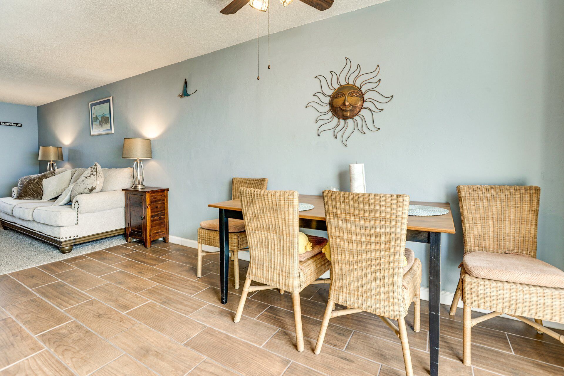 Ein Wohnzimmer mit einer Couch, zwei Beistelltischen, einem Esstisch mit Stühlen, einer Wanddekoration mit Sonnenelementen und einem Deckenventilator.