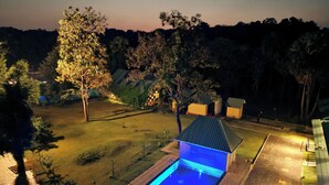 A rectangular swimming pool with a blue-lit interior, surrounded by a brick deck and a grassy area with trees. - Ibex Resorts Malampuzha Kava Eco Camp (Malampuzha-I)