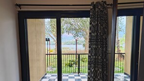 A room with a checkered floor, a view of a fenced outdoor area with greenery and a body of water, and a patterned curtain. - Ibex Resorts Malampuzha Kava Eco Camp (Malampuzha-I)