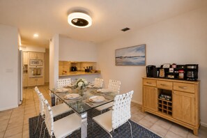 Een moderne keuken met een eethoek, met een glazen tafel, witte stoelen en een houten kast met keukenapparatuur.