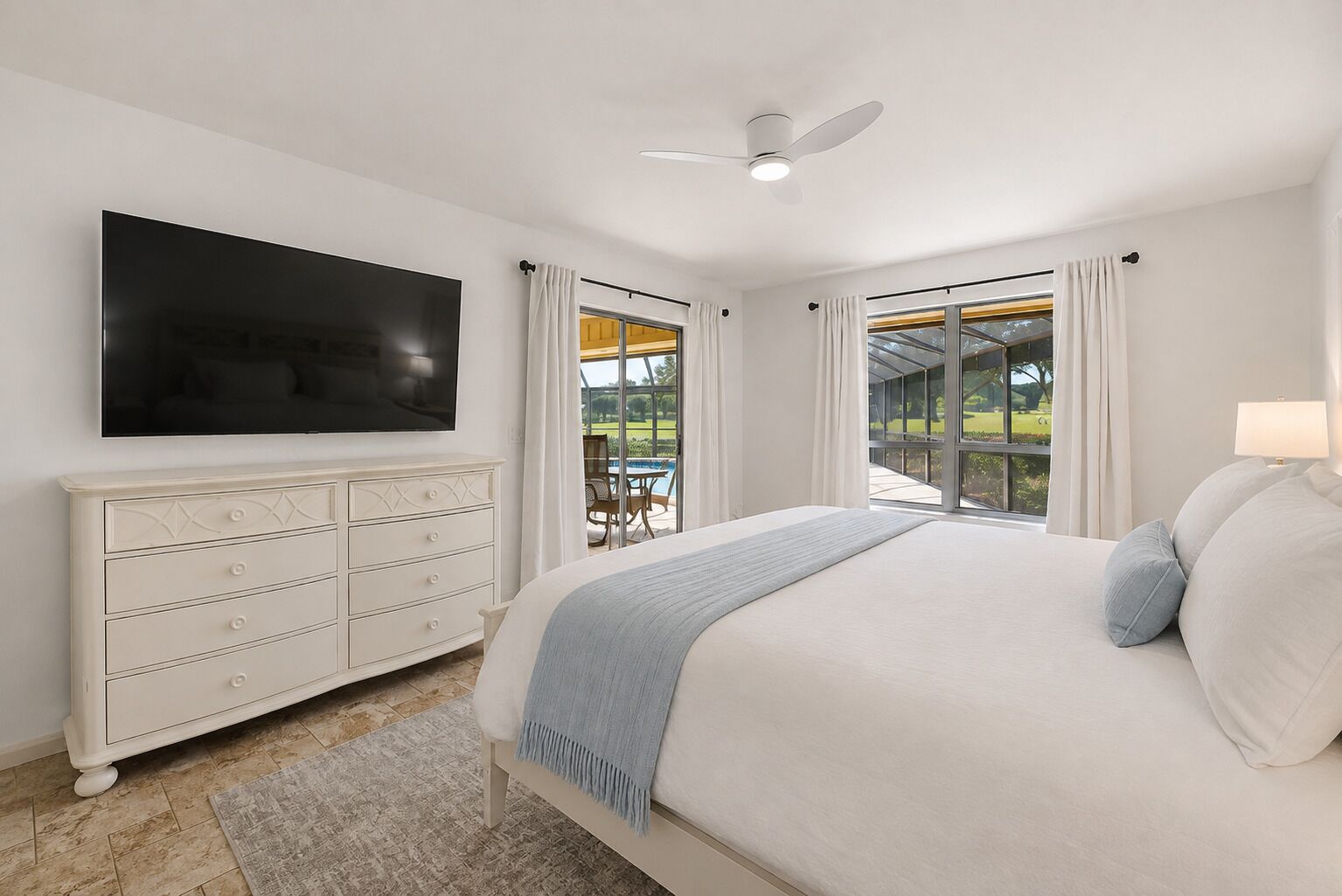 A bedroom with a large bed, a flat-screen TV, a dresser, and a view of a patio with outdoor furniture.