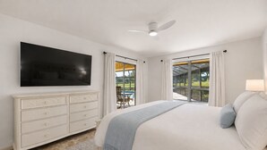 A bedroom with a large bed, a flat-screen TV, a dresser, and a view of a patio with outdoor furniture. - Greenside in Palm Beach Gardens - Private Pool & Golf Course Views! (Palm Beach Gardens)