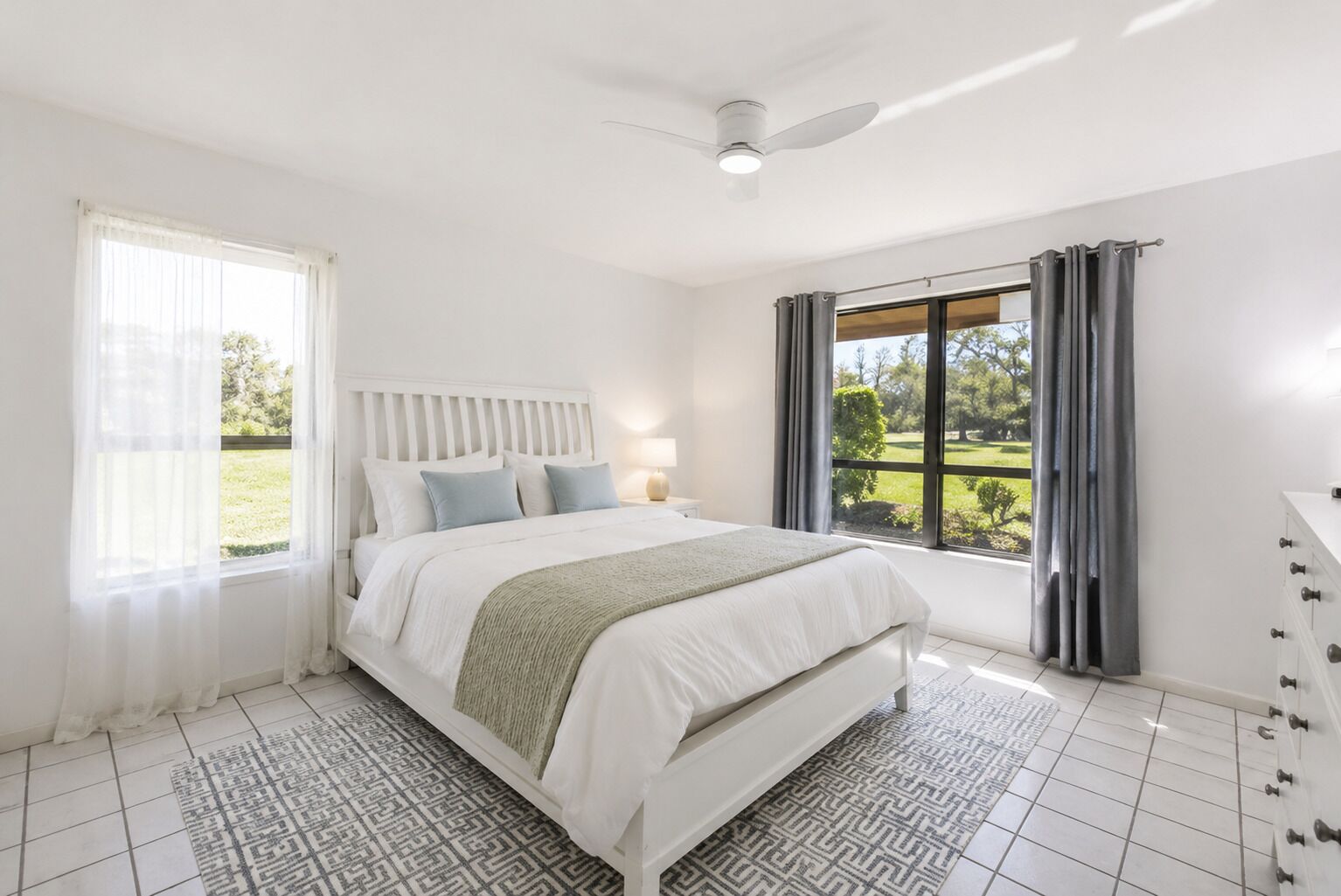 Un dormitorio con una cama, una mesita de noche, una lámpara, una ventana con cortinas y vistas al exterior.