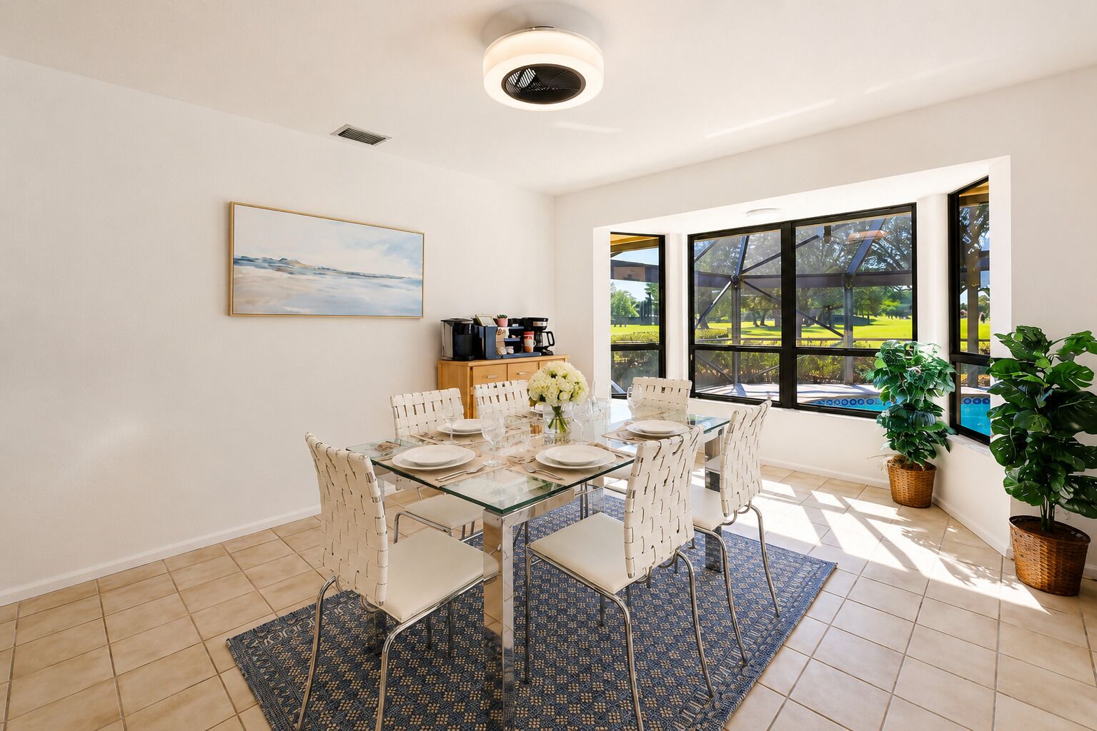 A dining area with a glass table, white chairs, a painting on the wall, and a view of a green landscape through large windows.