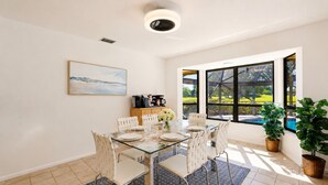 A dining area with a glass table, white chairs, a painting on the wall, and a view of a green landscape through large windows. - Greenside in Palm Beach Gardens - Private Pool & Golf Course Views! (Palm Beach Gardens)