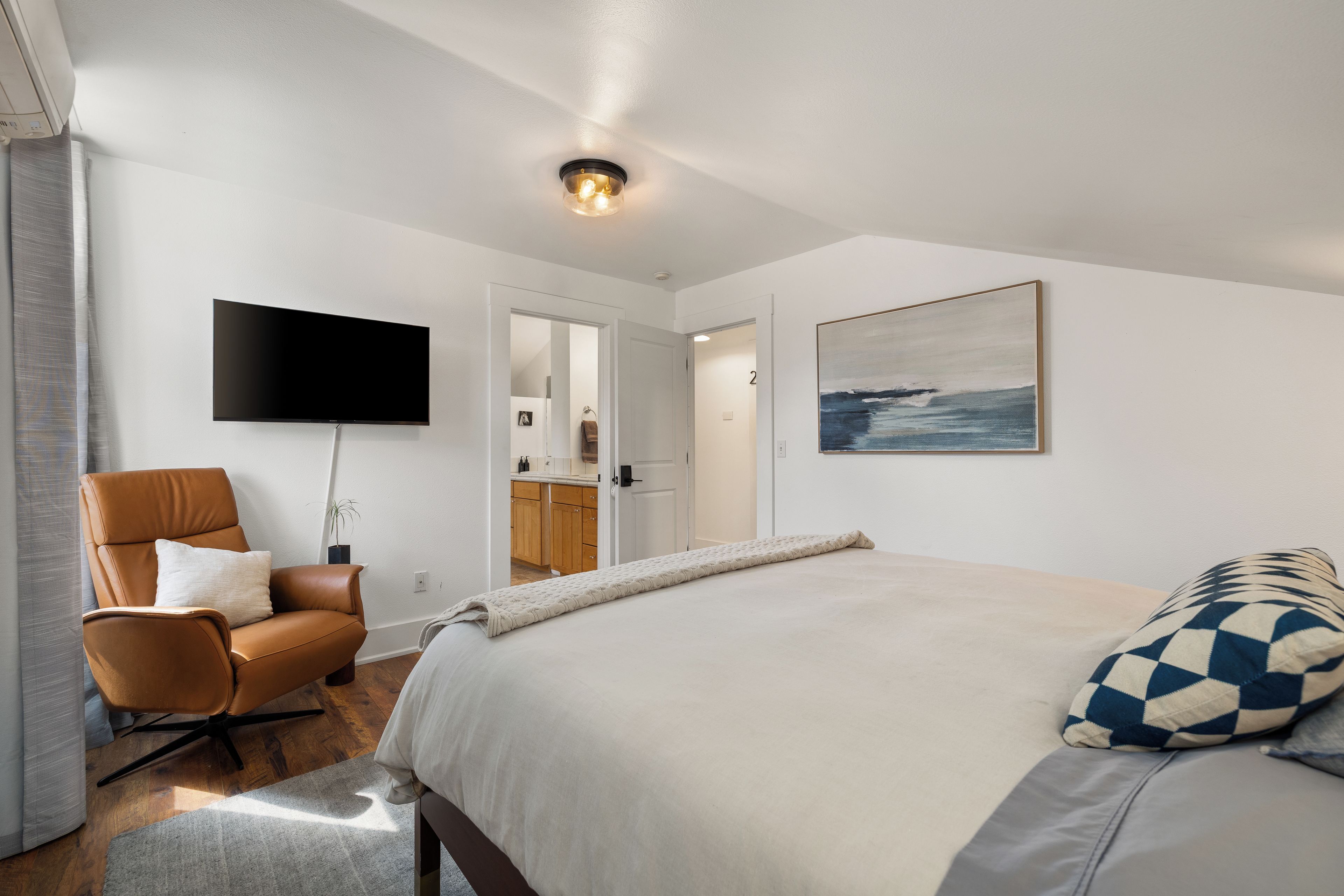 A bedroom with a bed, a chair, a flat-screen TV, a painting, and a door leading to a bathroom.