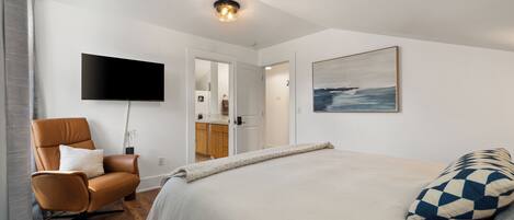 A bedroom with a bed, a chair, a flat-screen TV, a painting, and a door leading to a bathroom.