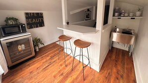A compact kitchen with a microwave, a refrigerator, wooden stools, a white countertop, and a shelf with kitchen items. - Spacious downtown loft, with soaking tub&gym access-walk to BSU and Boise River! (Boise)