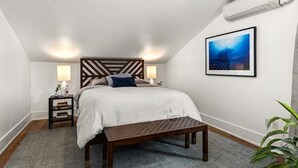A bedroom with a bed, bedside tables, a bench, a wall-mounted air conditioner, and a framed picture on the wall. - Spacious downtown loft, with soaking tub&gym access-walk to BSU and Boise River! (Boise)