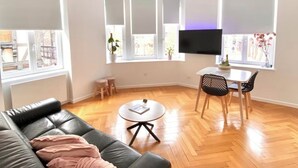 Ein modernes Wohnzimmer mit einem schwarzen Ledersofa, einem runden Couchtisch, einem Esstisch mit Stühlen und großen Fenstern mit Jalousien.