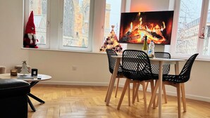 Ein modernes Wohnzimmer mit Holzdielen im Fischgrätmuster, einem Essbereich mit Fernseher, der einen Kamin zeigt, und großen Fenstern mit Blick auf ein Gebäude.