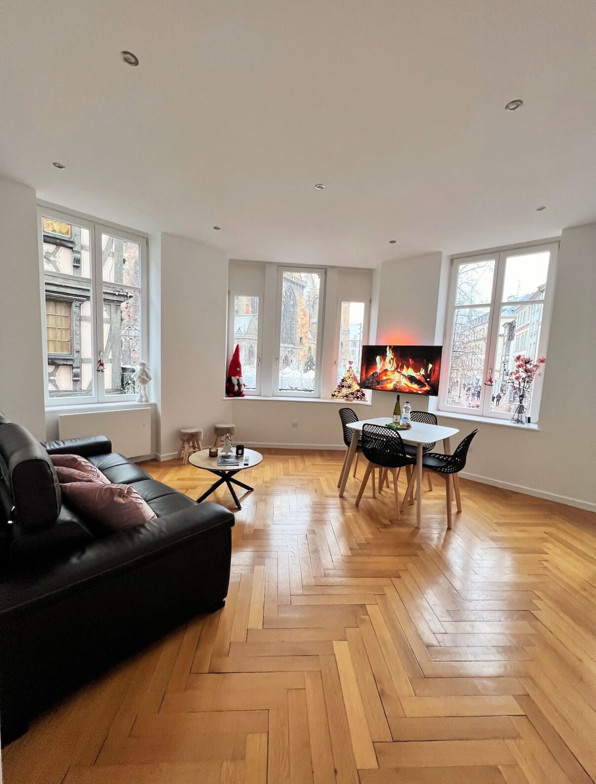 Een moderne woonkamer met een zwarte leren bank, een ronde salontafel en een eethoek met een witte tafel en zwarte stoelen. Grote ramen bieden uitzicht op de stad.