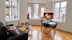 Ein modernes Wohnzimmer mit einer schwarzen Ledersofa, einem runden Couchtisch und einem Essbereich mit weißem Tisch und schwarzen Stühlen. Große Fenster bieten Blick auf die Stadt.