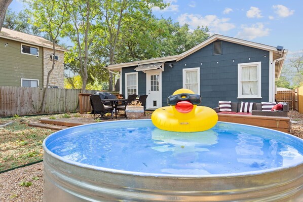 Un patio trasero con piscina, un pato inflable y una casa con terraza y mobiliario de exterior.