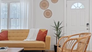 Een woonkamer met een geel sofa, een rieten stoel, een bloempot en een witte deur.
