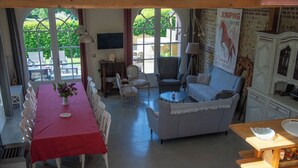 Un comedor con mantel rojo, sillas blancas y un sofá. Hay un cartel en la pared y una ventana con vistas a zonas verdes.