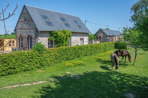 Ein Steinhaus mit geneigtem Dach und Oberlichtern, umgeben von einer gepflegten Hecke und einer Rasenfläche mit Pferden.