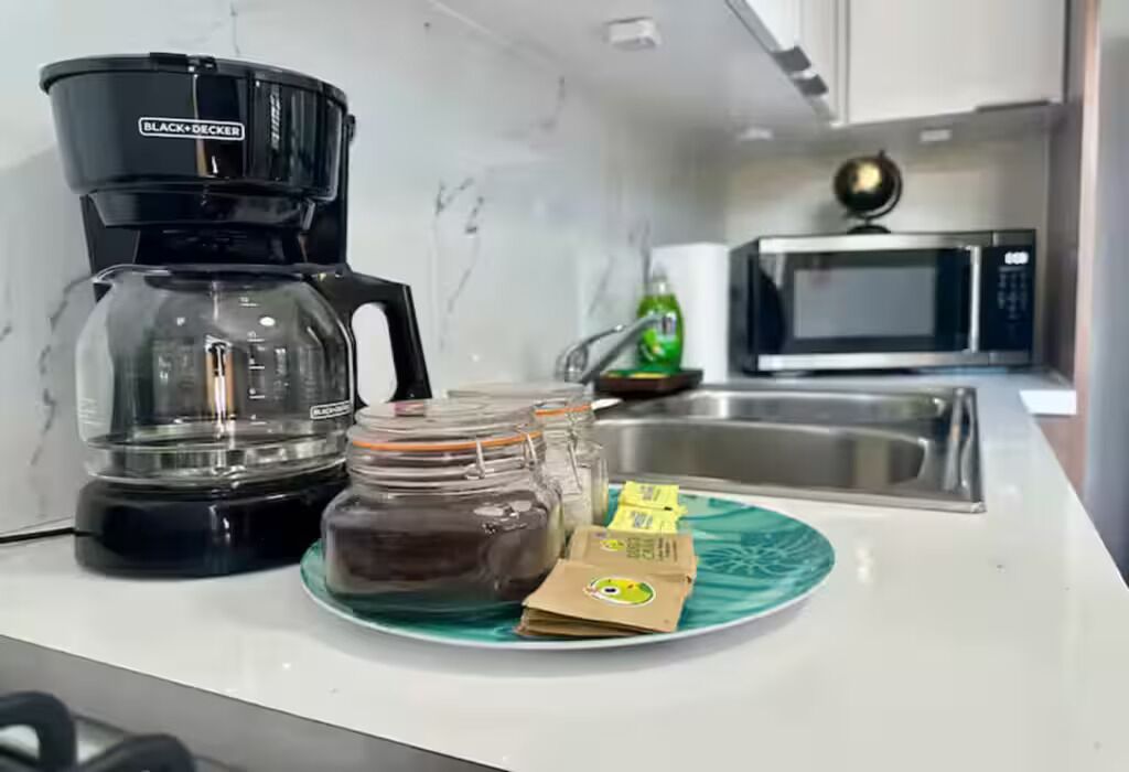 Une cuisine équipée d’une machine à café noire, d’un four à micro-ondes, d’un évier, ainsi que d’une assiette verte contenant des aliments et une dosette de café.
