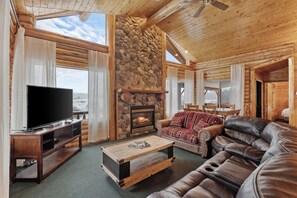 A cozy log cabin interior with a stone fireplace, leather sofas, a flat-screen TV, and large windows offering a view of the outdoors. - Henry`s Fork Cabin | Steps to River, Hot Tub, Amazing Views, + BBQ! (Island Park)
