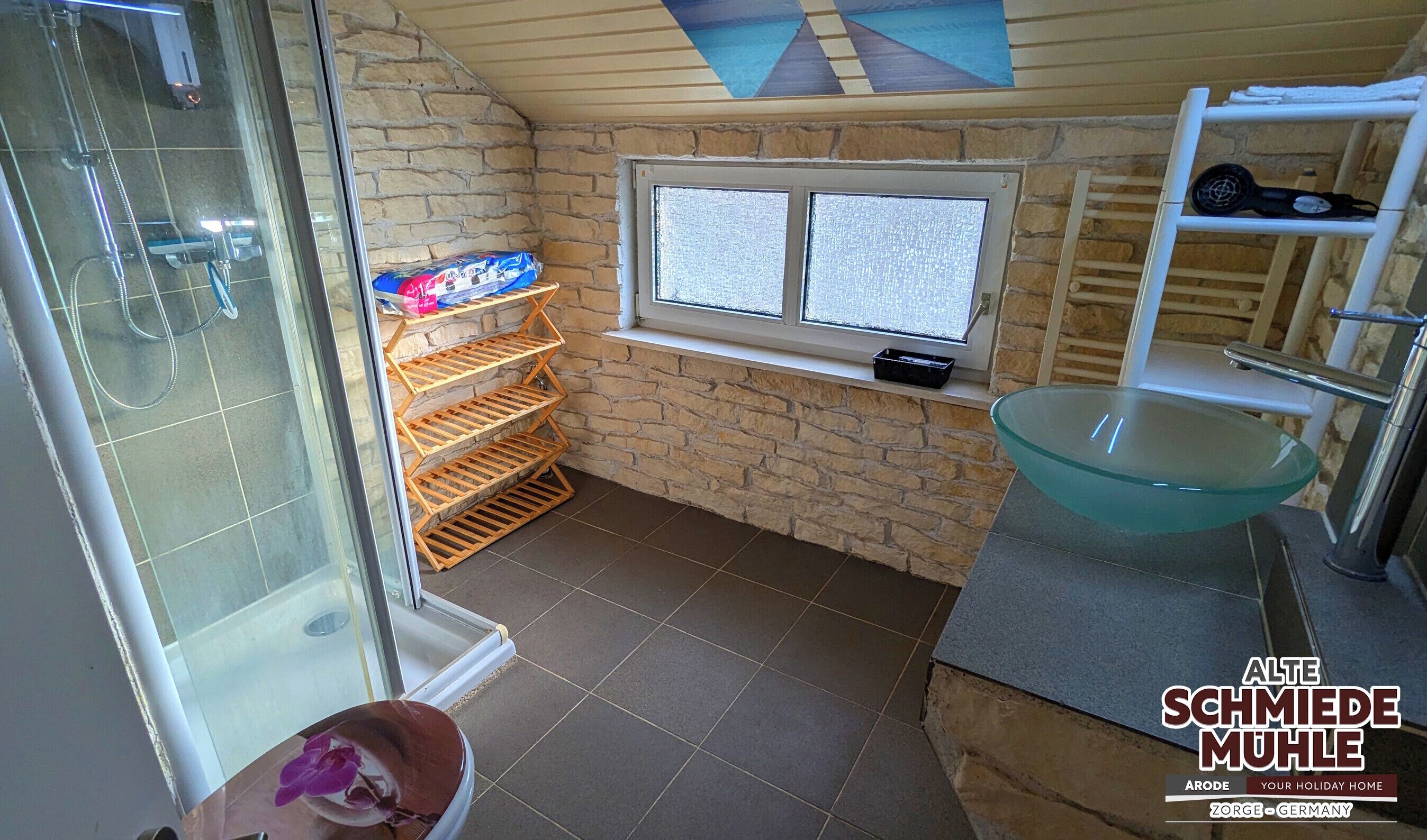 Une salle de bain avec une douche en verre, une étagère en bois où sont rangés les articles de toilette, une fenêtre et un lavabo en verre.
