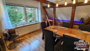 Ein Essbereich mit einem Holztisch, gedeckt für sechs Personen, umgeben von schwarzen Stühlen. Zwei Fenster mit Vorhängen, ein Heizkörper darunter und eine Holzbalkenkonstruktion über dem Tisch.