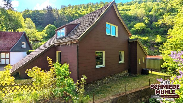 Ein traditionelles Holzhaus mit steilem Dach, eingebettet in eine üppige, grüne Waldlandschaft.