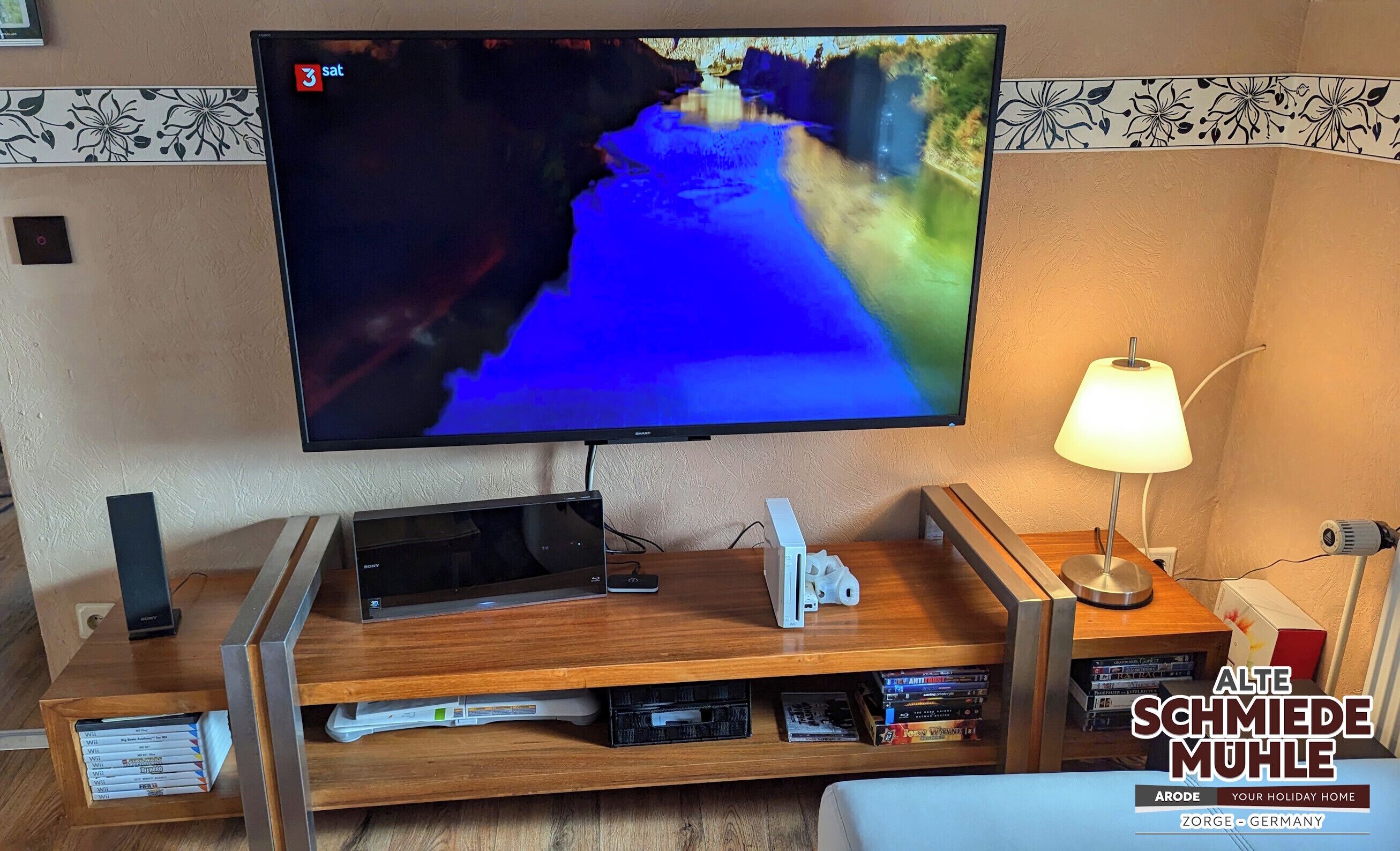 Un televisor de pantalla plana montado en la pared, un mueble de madera con una lámpara y un estante con DVD.