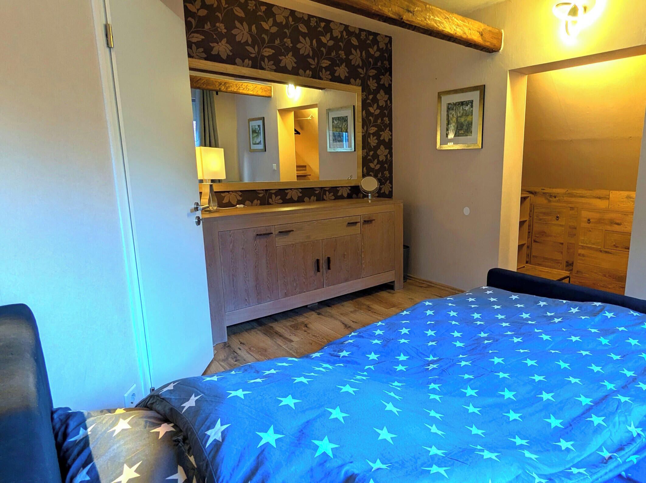 A bedroom with a bed, a wooden dresser, a mirror, and a door leading to another room.