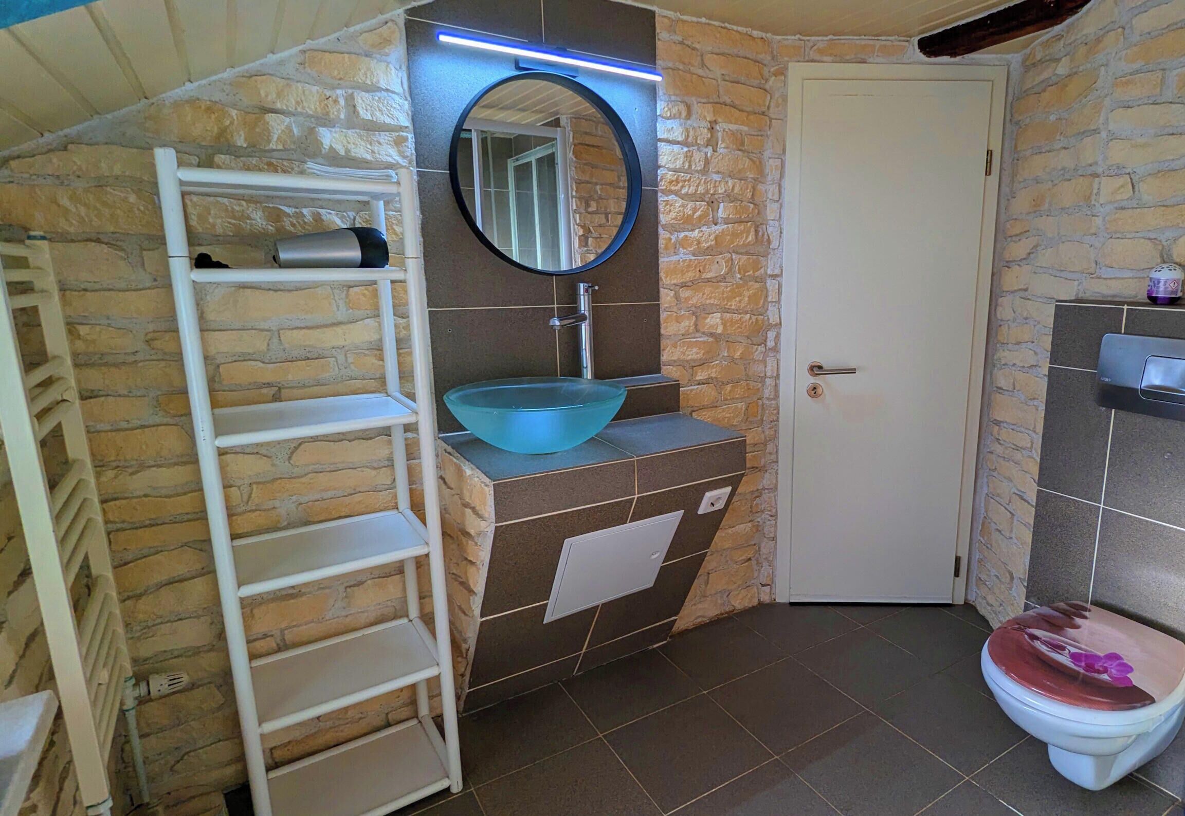 Une salle de bain avec un mur en pierre, un lavabo bleu, une porte blanche et des toilettes avec un abattant rose.