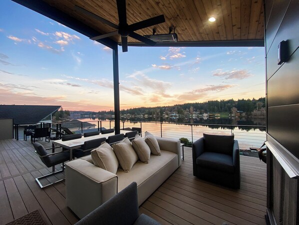 Une terrasse avec un canapé, des chaises et une table, offrant une vue sur un lac et un magnifique coucher de soleil.