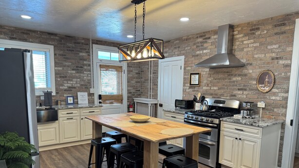 A modern kitchen with a wooden island, black bar stools, a stainless steel stove, and a brick wall. - Beautiful Civil War home in Downtown Gettysburg! (Gettysburg)