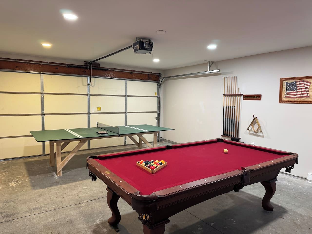 A billiards table with balls, a green table tennis table, and a ping pong table in a room with a garage door and a flag on the wall.