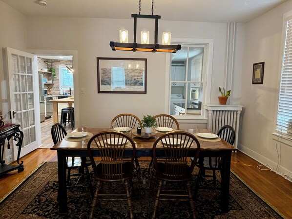 Comedor con mesa de madera, cuatro sillas, una araña, un cuadro enmarcado y vista a la cocina.