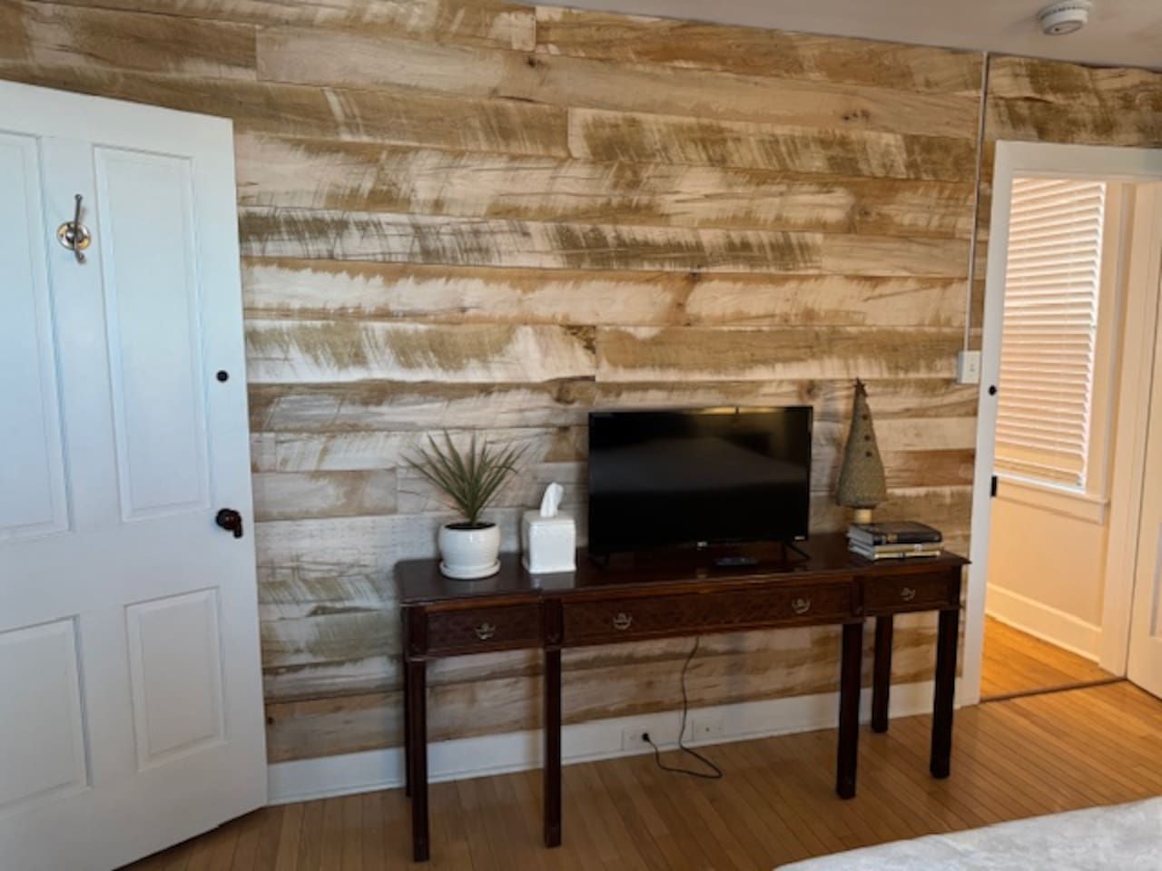 Ein Zimmer mit einer Holzwand, einer weißen Tür, einem dunklen Holz-Konsolentisch mit Fernseher, einer kleinen Pflanze und einer Lampe.