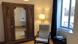Quarto com guarda-roupa de madeira, uma cadeira, uma mesinha, uma lâmpada e uma janela com vista.
