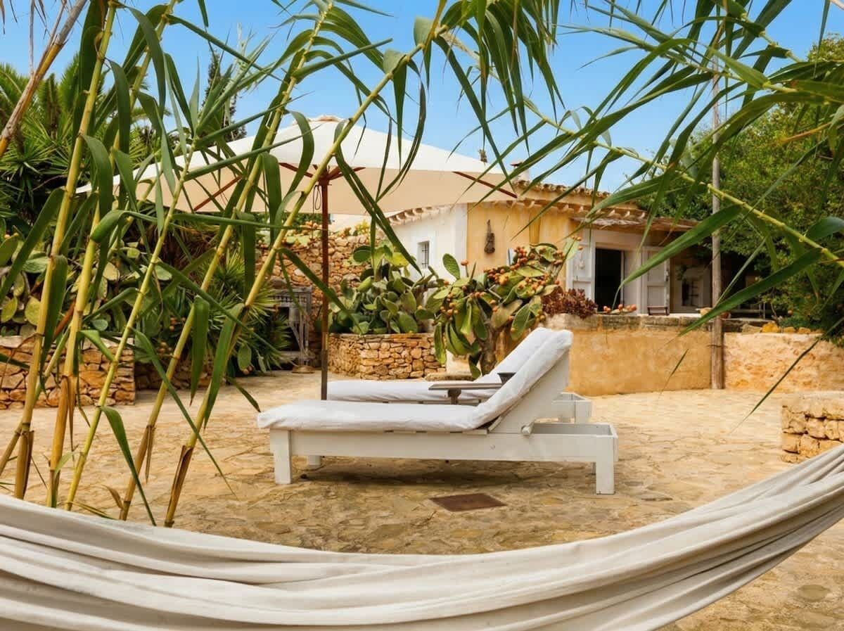 A hammock with a white lounge chair, a stone wall, and a building with a tiled roof in the background.
