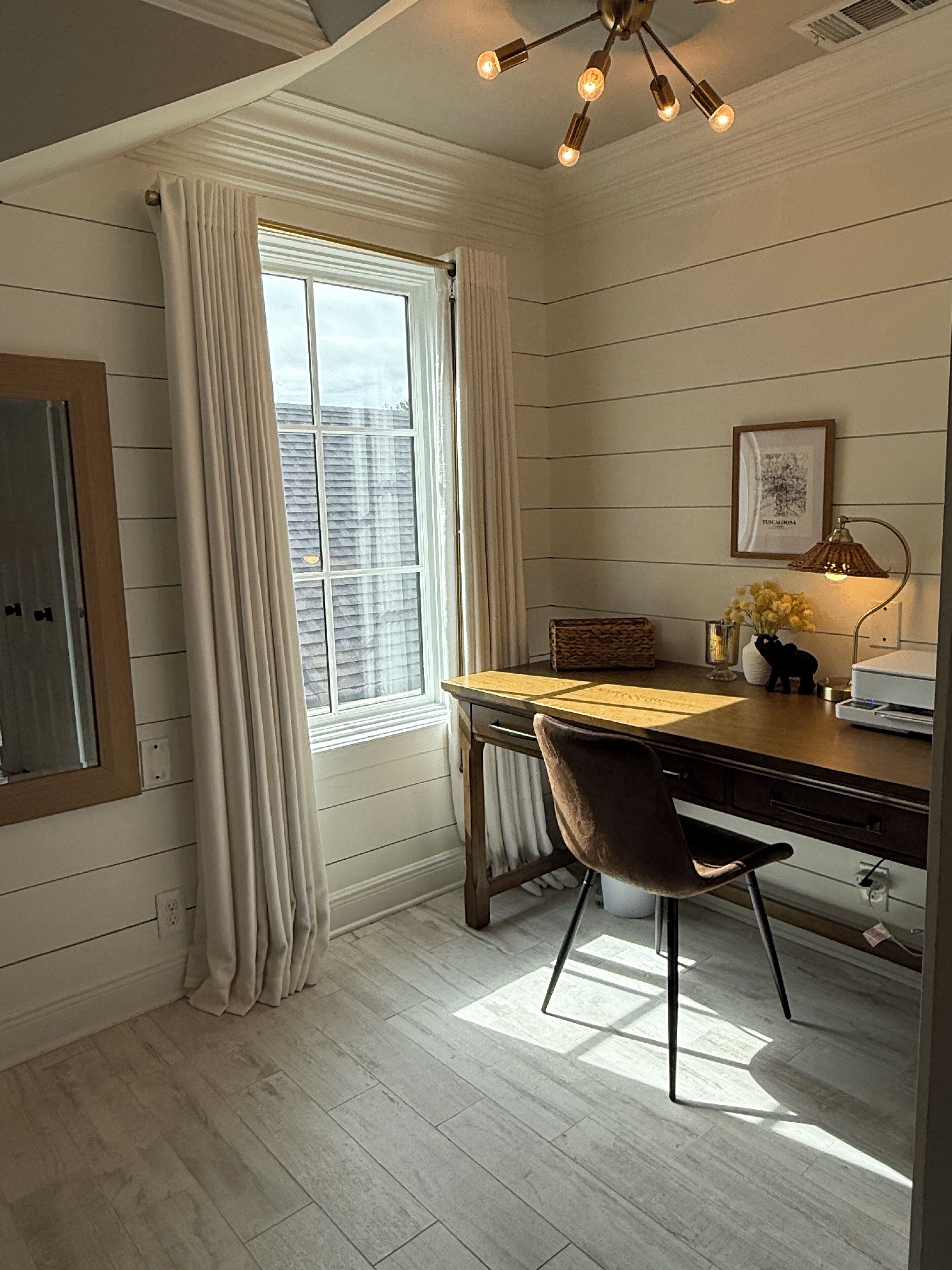 A room with a wooden desk, a chair, a window with curtains, a wall-mounted lamp, and a framed picture.