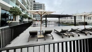 Área de piscina no terraço com espreguiçadeiras e guarda-sóis, cercada por edifícios modernos.