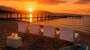 Ein Strand mit Liegestühlen und einem Stand-Up-Paddleboard, im Hintergrund ein Sonnenuntergang und ein Steg, der sich ins Wasser erstreckt.