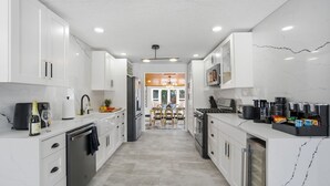 A modern kitchen with white cabinetry, a central island, and a dining area in the background. - King Bed w/ Game Room near Massmutual w/ Riverview (Chicopee)