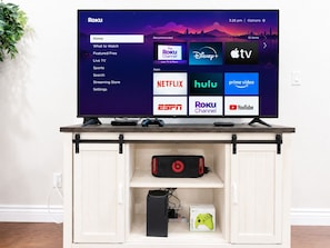 Ein Roku-Fernseher auf einer weißen Unterhaltungsanlage mit einem Spiel-Controller und einem PlayStation-Controller.