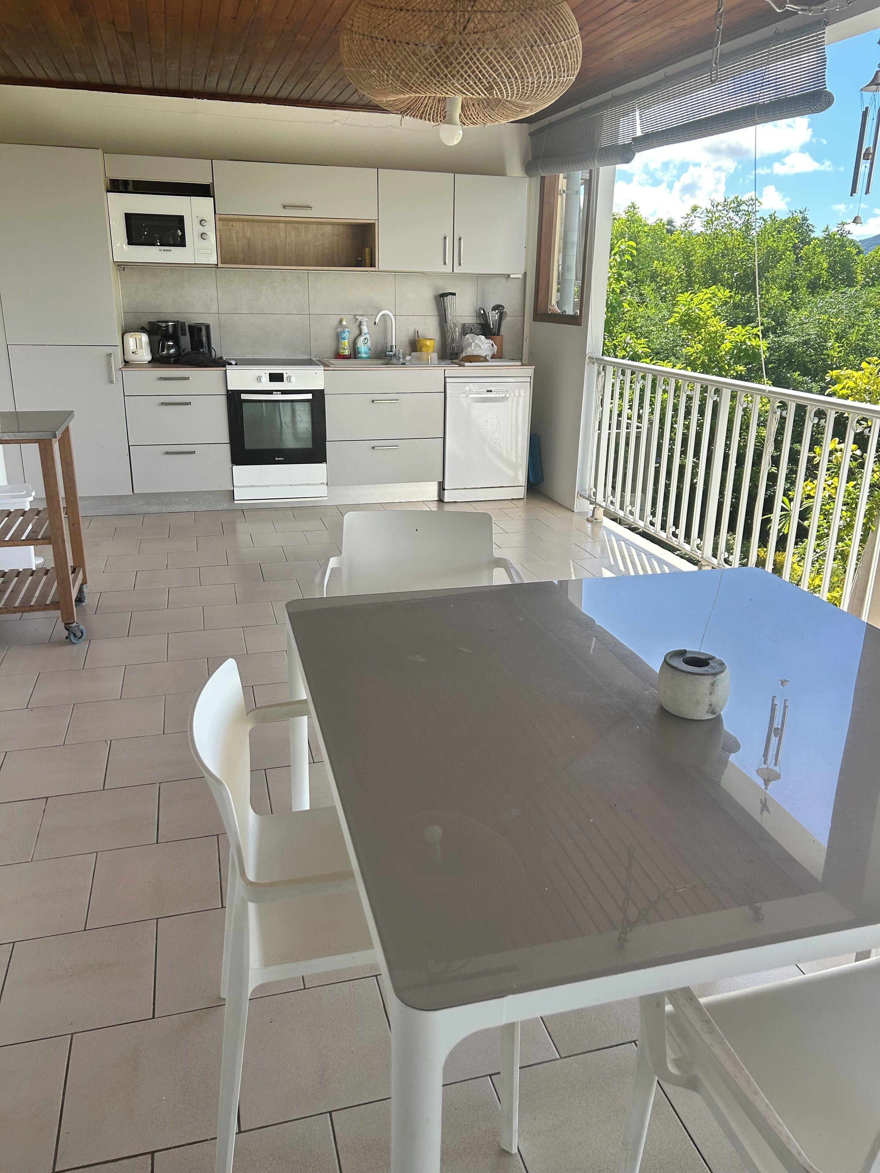 Een moderne keuken met witte kastjes, een magnetron, fornuis en vaatwasser. Er staat een eettafel met stoelen en er is uitzicht op groen via een balkonhek.