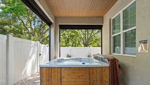 Een jacuzzi in een overdekte buitenruimte met een houten terras, een witte omheining en een raam met zonwering.