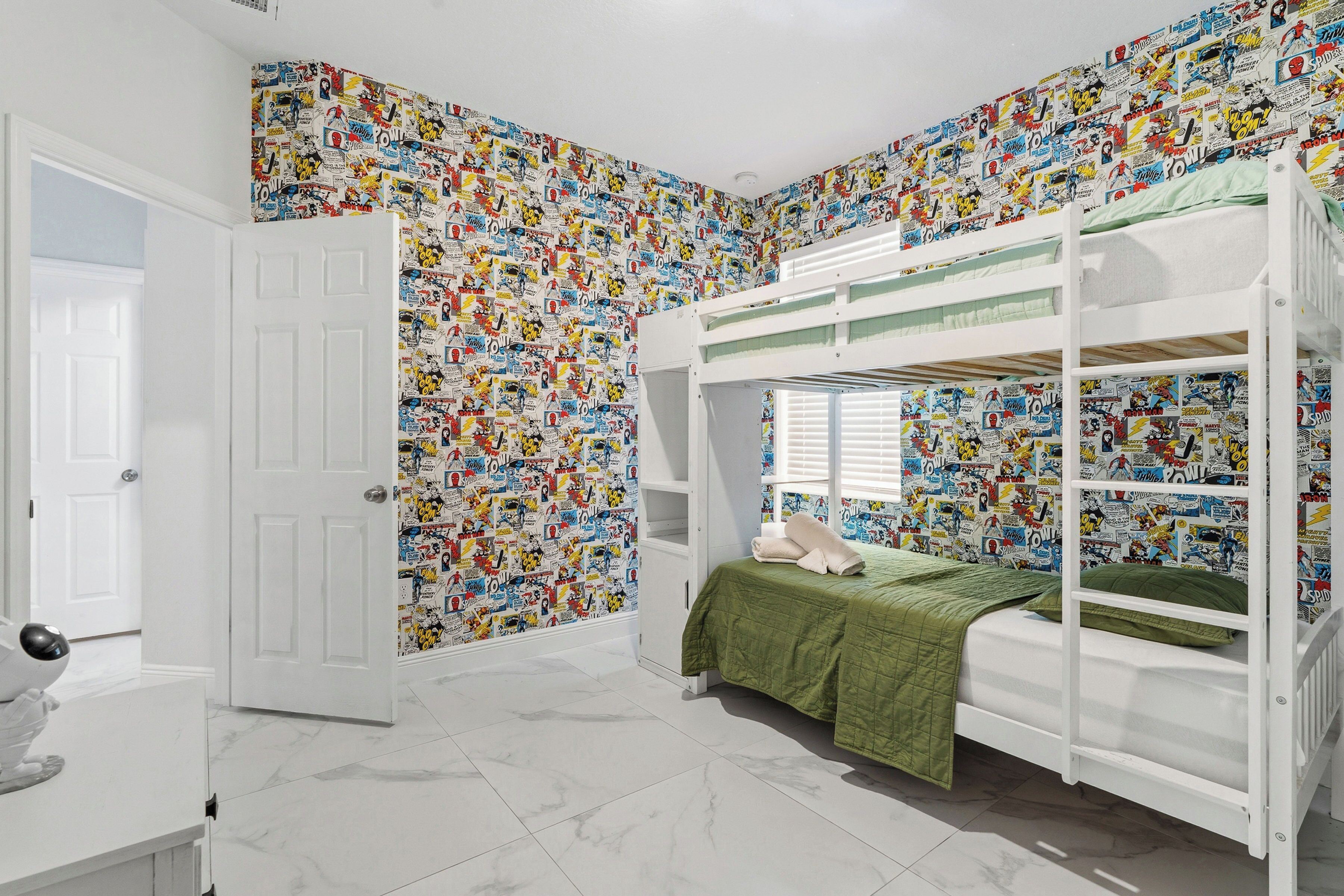 A room with a bunk bed, a door, a white dresser, and a wall covered with comic book-themed wallpaper.
