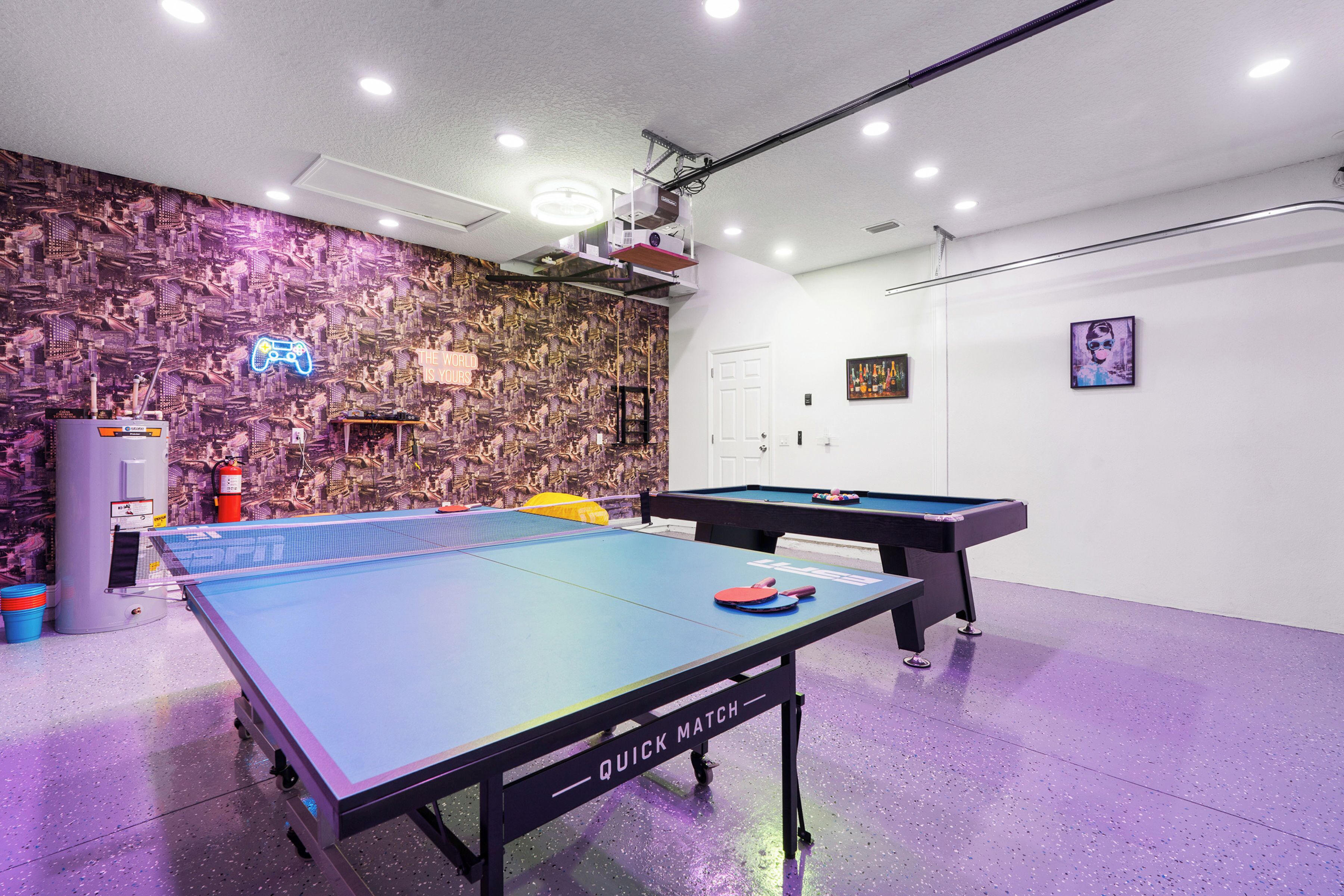 Une salle de billard avec deux tables, un panneau lumineux représentant une manette de jeu et un projecteur fixé au plafond.
