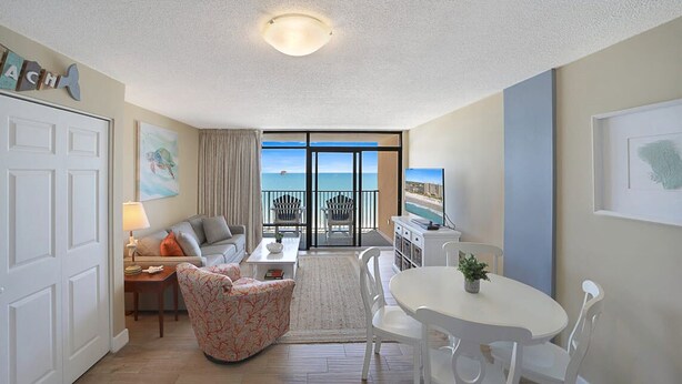 A spacious living room with a sofa, armchair, and dining table. A large window offers a view of the ocean. - Perfect 1-bedroom ocean front condo in charming North Myrtle Beach sleeps 6 (North Myrtle Beach)