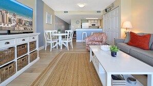 A living room with a television, white furniture, a kitchen in the background, and a view of the cityscape. - Perfect 1-bedroom ocean front condo in charming North Myrtle Beach sleeps 6 (North Myrtle Beach)