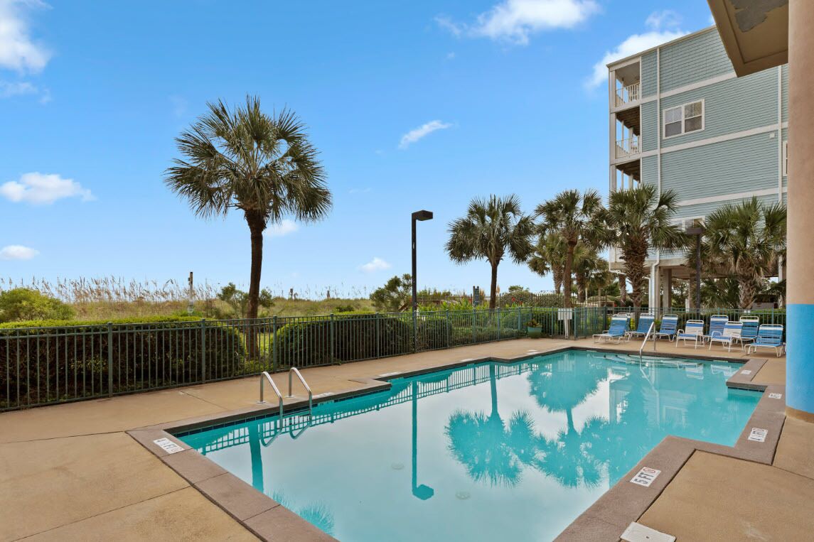 A pool area with clear blue water, surrounded by a fence and palm trees.