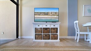 A room with a television displaying a beach view, a white cabinet with wicker baskets, a white chair, and a dining table. - Perfect 1-bedroom ocean front condo in charming North Myrtle Beach sleeps 6 (North Myrtle Beach)