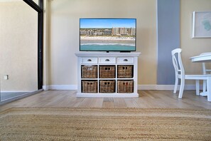 A room with a television displaying a beach view, a white cabinet with wicker baskets, a white chair, and a dining table. - Perfect 1-bedroom ocean front condo in charming North Myrtle Beach sleeps 6 (North Myrtle Beach)